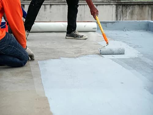 Two workers roller-applying a grey waterproof membrane across a concrete terrace