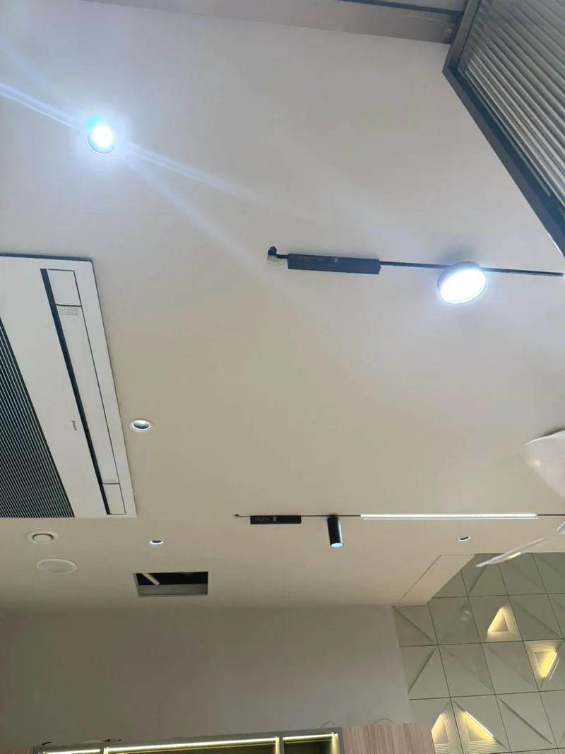 Ceiling with recessed downlights, a black magnetic track light run, a cassette air-conditioning unit and a faceted panel section